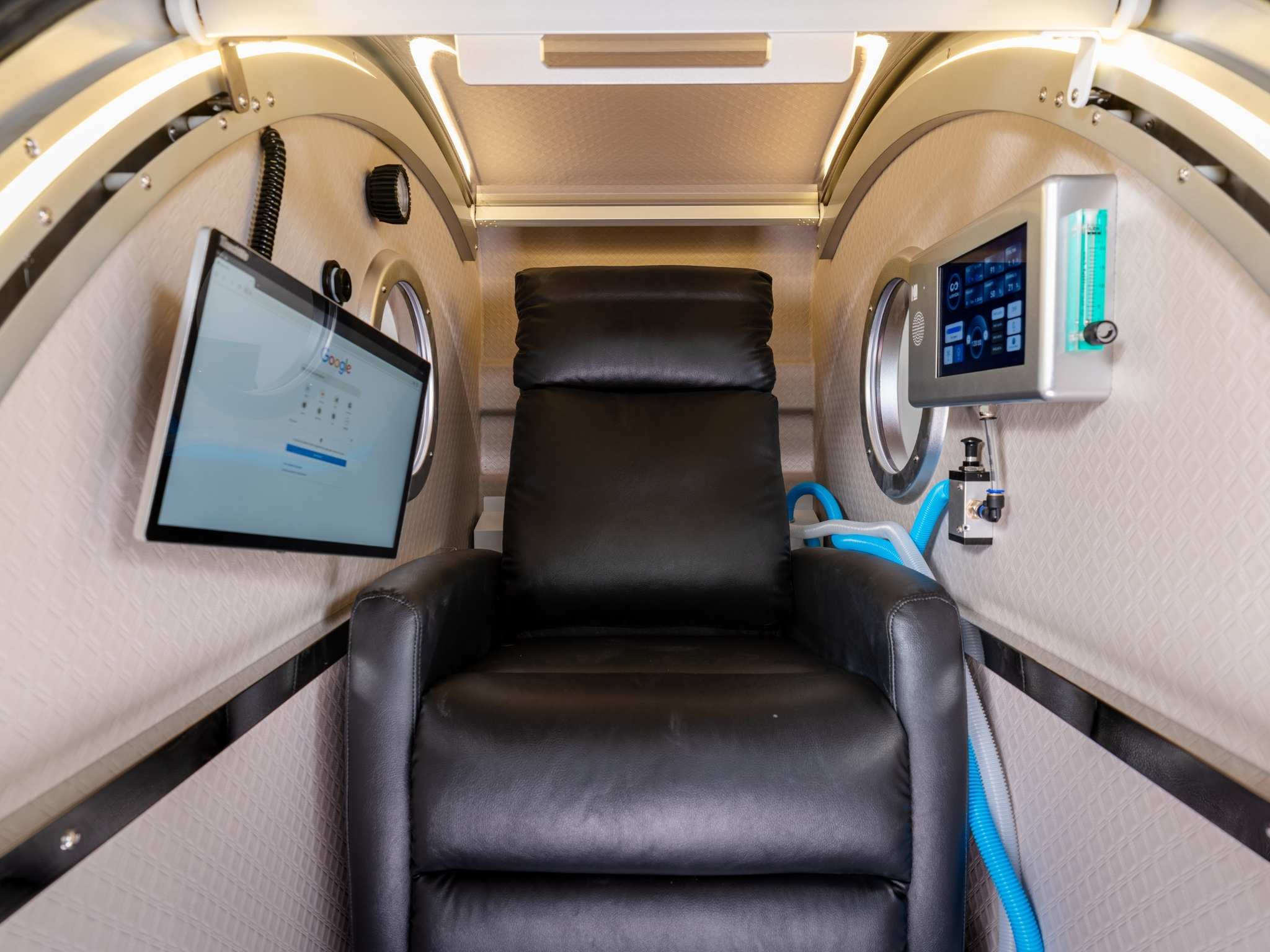 Interior of a seated hyperbaric oxygen chamber featuring a reclining leather chair, touchscreen controls, oxygen monitor, and display screen.