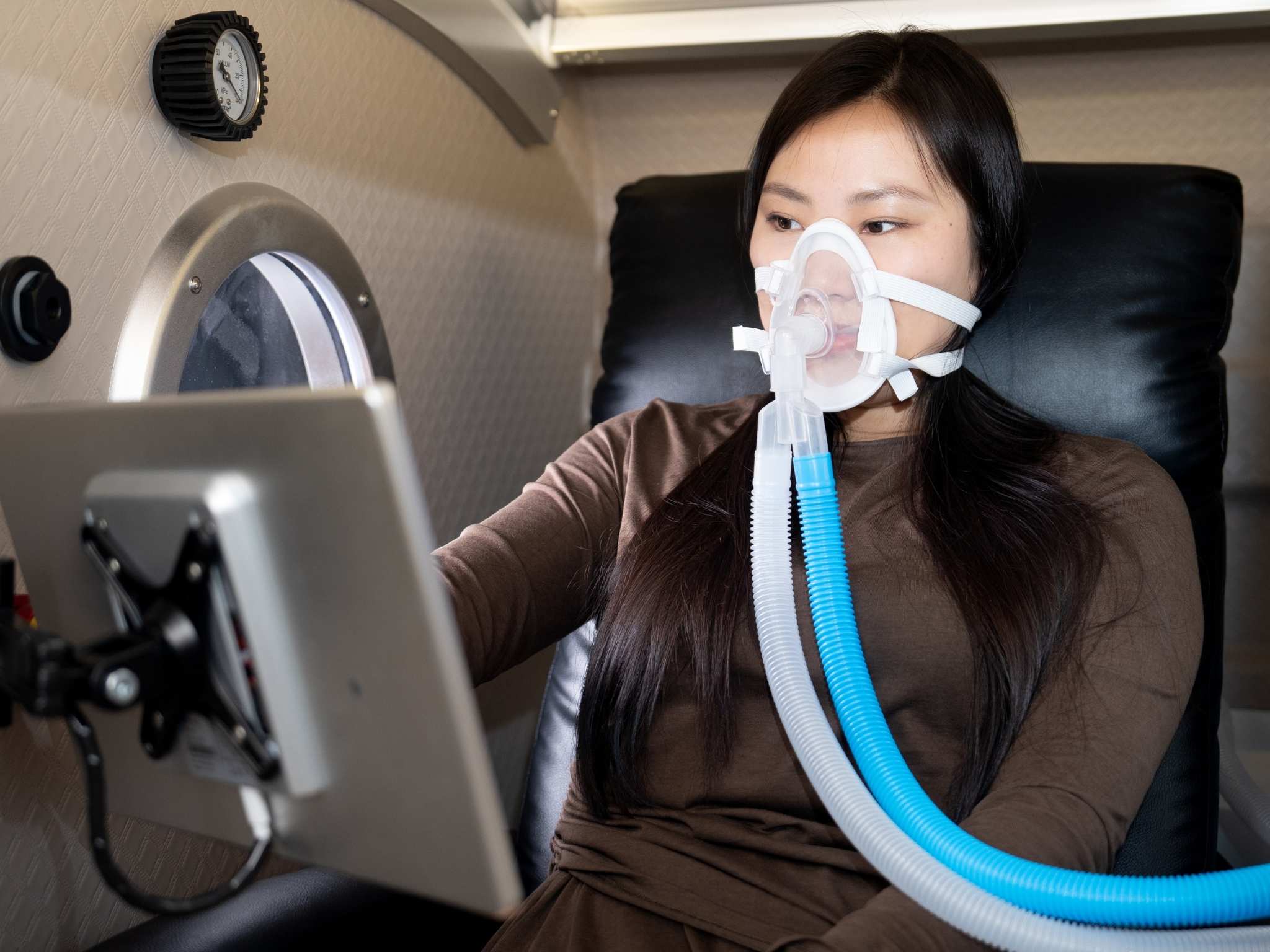 Person wearing an oxygen mask sits comfortably inside a seated hyperbaric oxygen chamber while using a mounted touchscreen display.