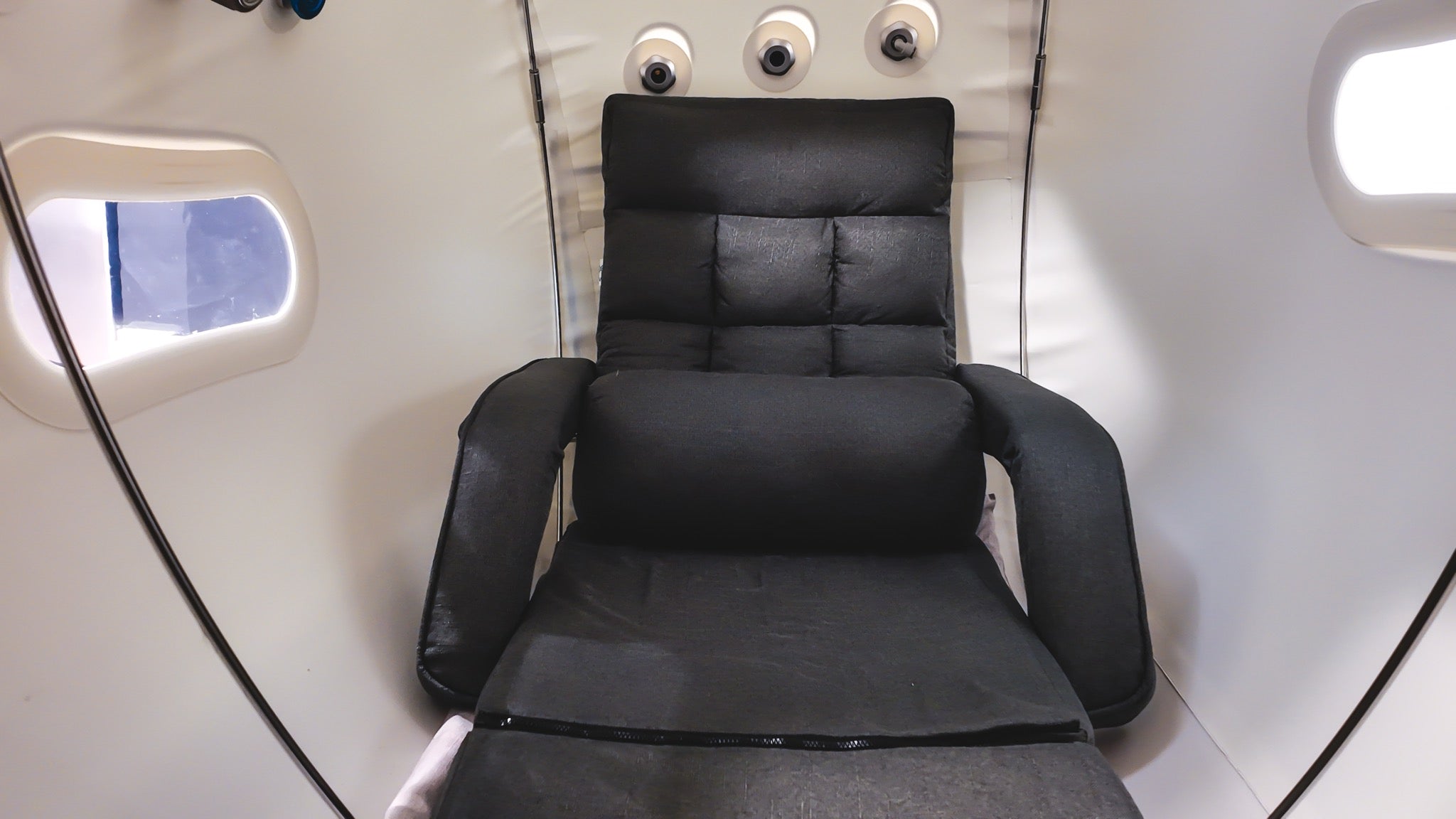 Interior of an AIRVIDA hyperbaric chamber featuring a padded black reclining chair, viewing windows, air ports, and a spacious white cabin.