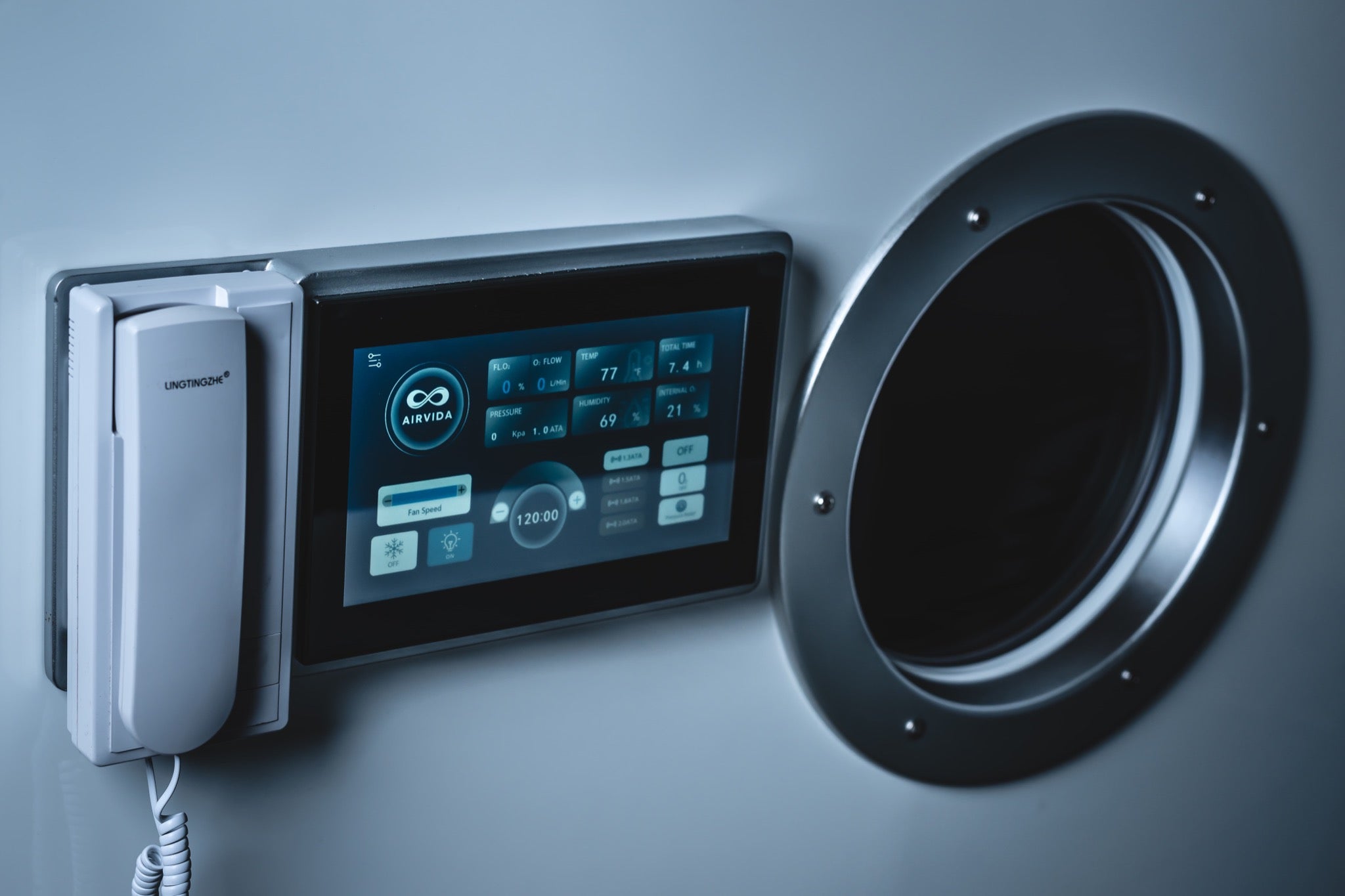 Close-up of an Airvida hyperbaric chamber touchscreen and communication handset beside a circular viewing window.