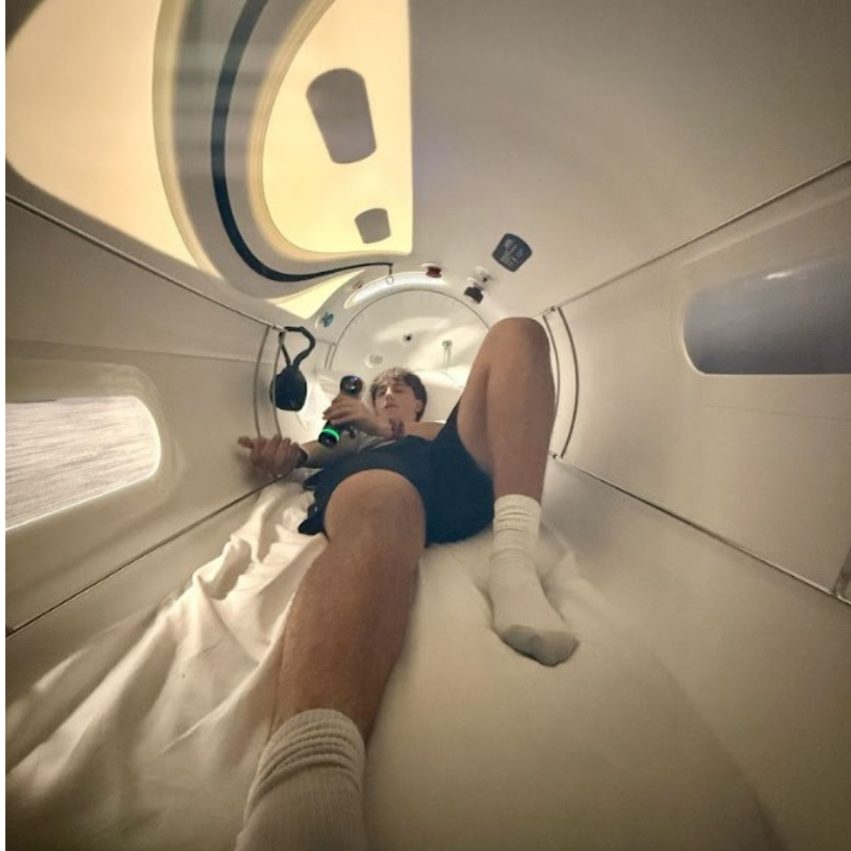 A person is lying inside a bright, narrow white hyperbaric chamber.
