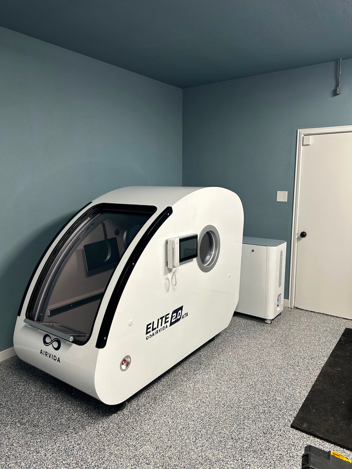 White Airvida Elite 2.0 ATA hyperbaric chamber with clear curved door, digital controls and oxygen unit installed in a compact blue-walled wellness room.
