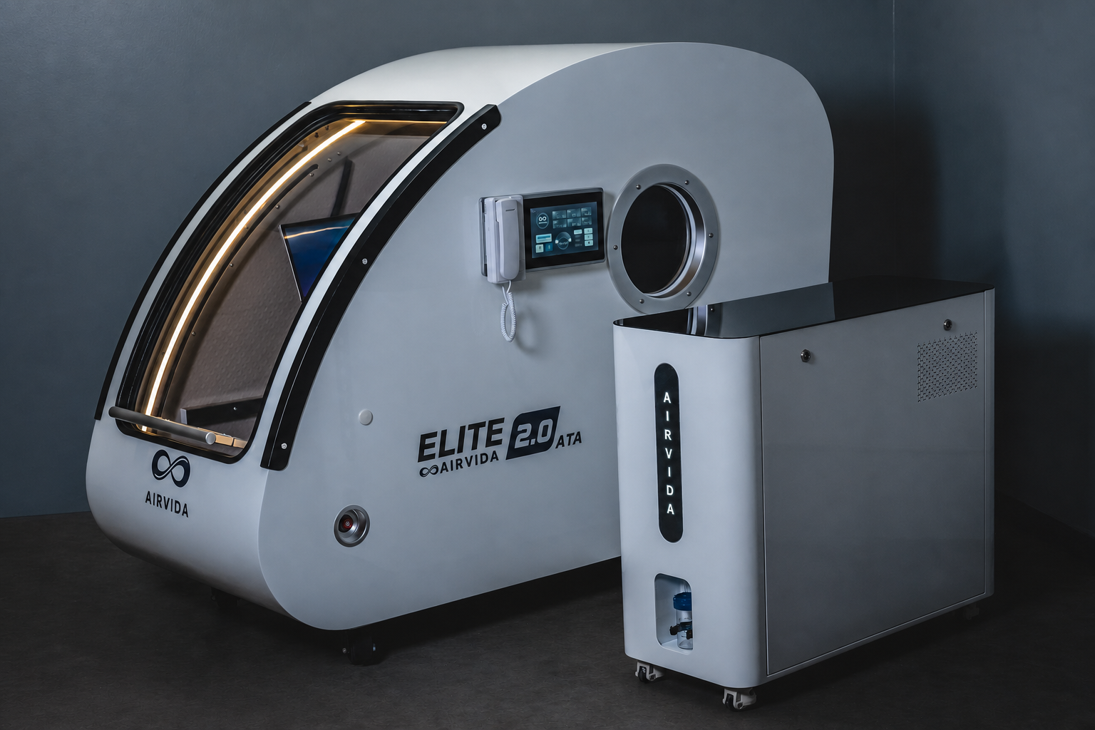 A white AIRVIDA Elite 2.0 Hyperbaric machine with a transparent curved viewing window, shown alongside its white auxiliary air supply unit against a dark gray studio background.