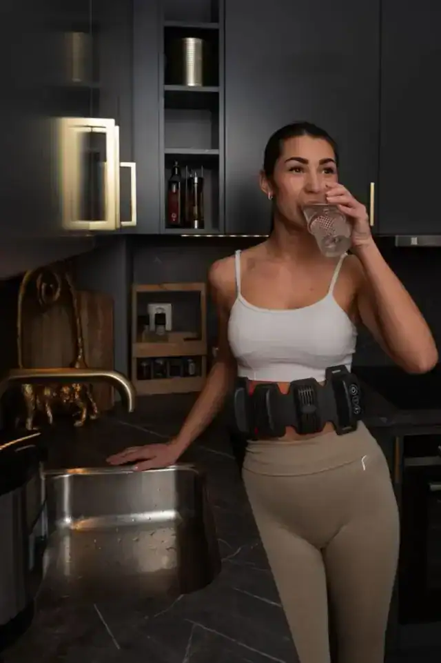 Woman drinking water in a kitchen wearing a FlexBeam Portable Red Light Therapy Device around her waist.