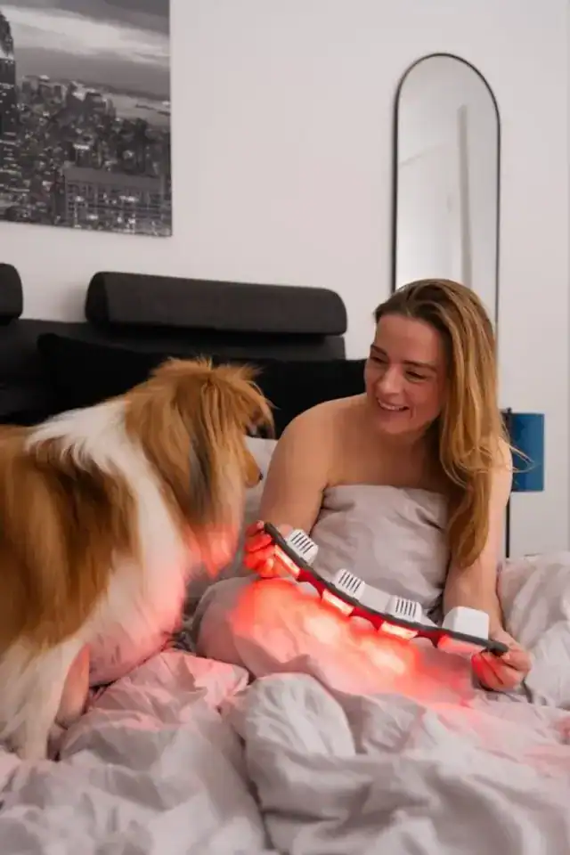 Woman using a FlexBeam Portable Red Light Therapy Device on her bed with a dog beside her.