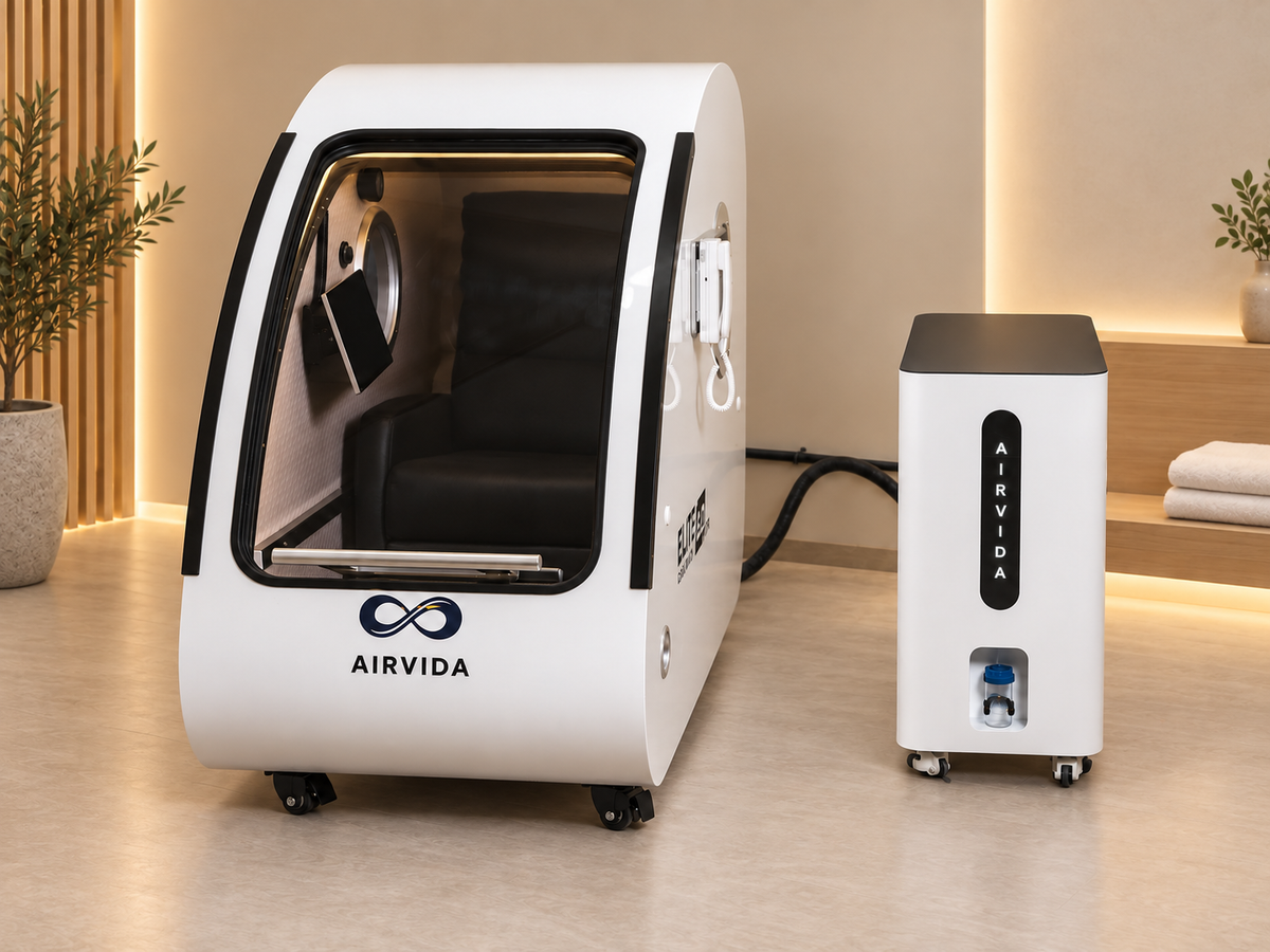 White Airvida hyperbaric chamber with clear curved door, cushioned interior and matching oxygen unit on wheels displayed in a warm modern wellness room setting.