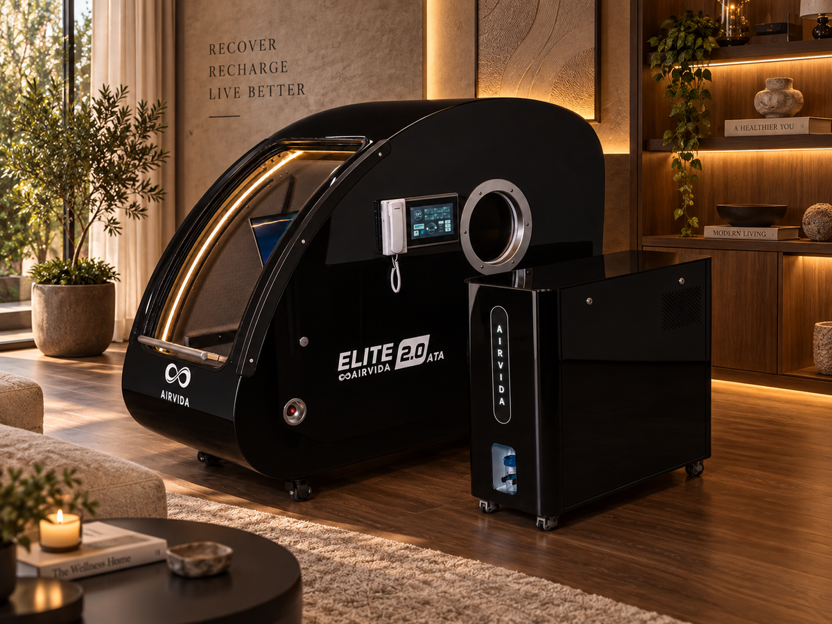 Black Airvida Elite 2.0 hyperbaric chamber with control unit in a luxurious home wellness room featuring warm wood, plants, shelving, and soft lighting.