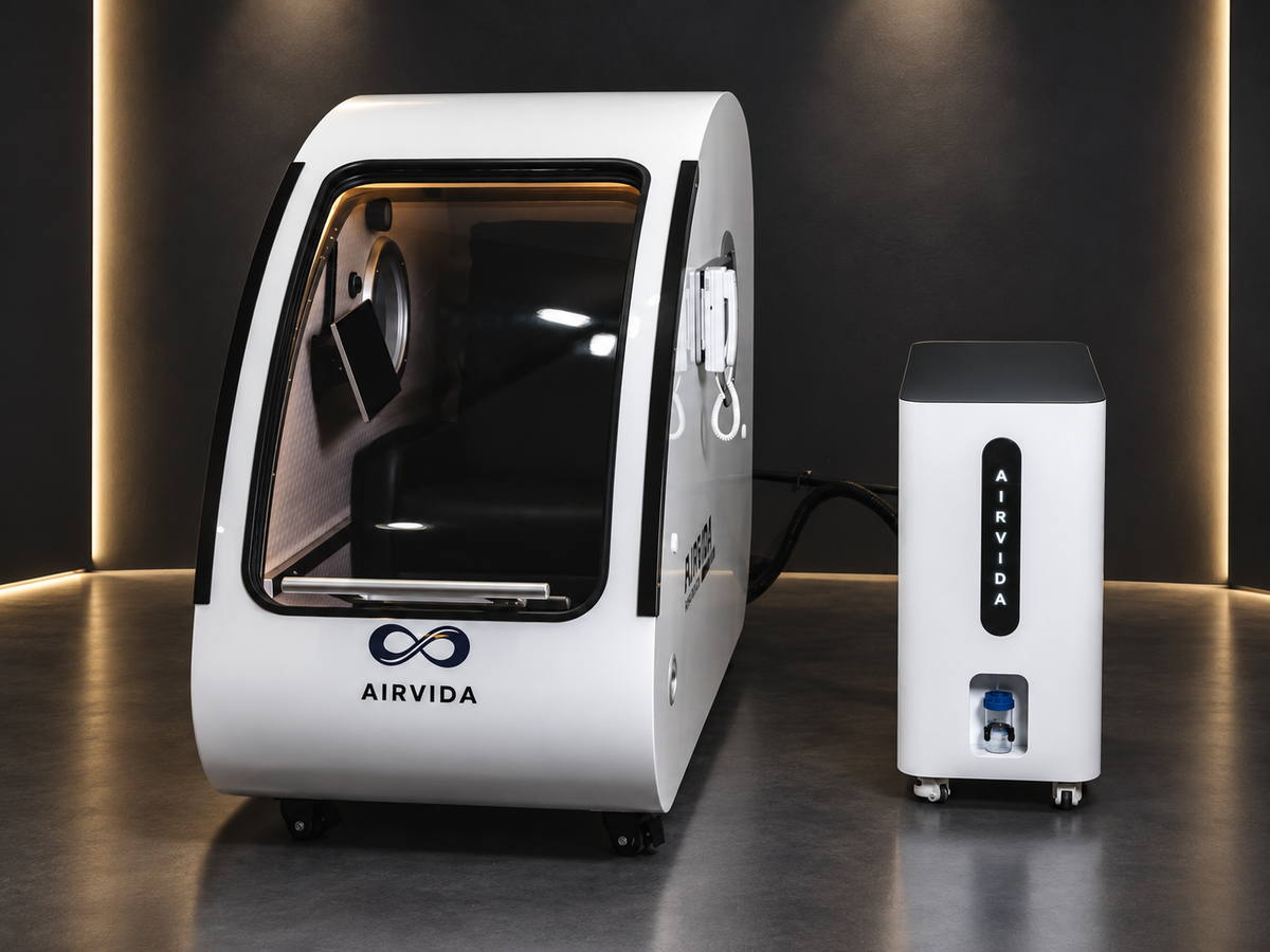 White Airvida hyperbaric chamber with clear curved door and matching oxygen unit on wheels, displayed against a dark wall with warm modern accent lighting.