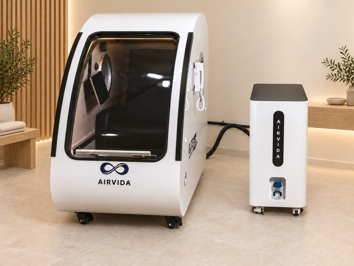 White Airvida hyperbaric chamber with external oxygen system displayed in a modern wellness room with warm wood accents, soft lighting, plants, and neutral decor.