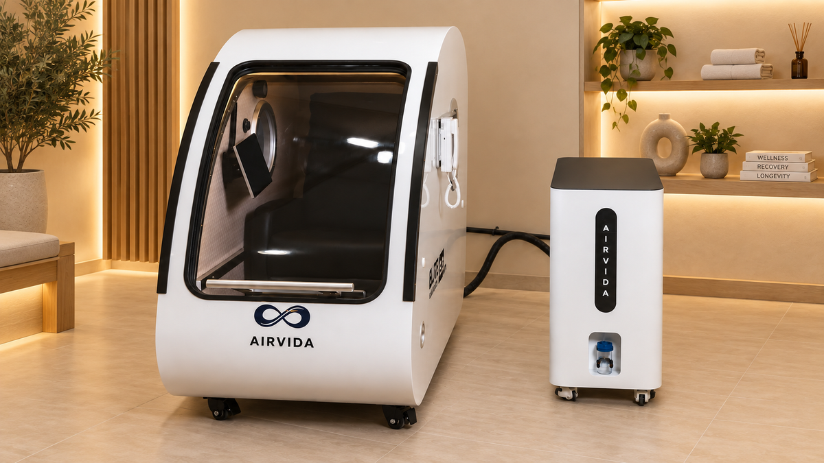 White Airvida hyperbaric chamber with oxygen system in a warm modern wellness room featuring wood accents, plants, towels, and softly illuminated shelves.