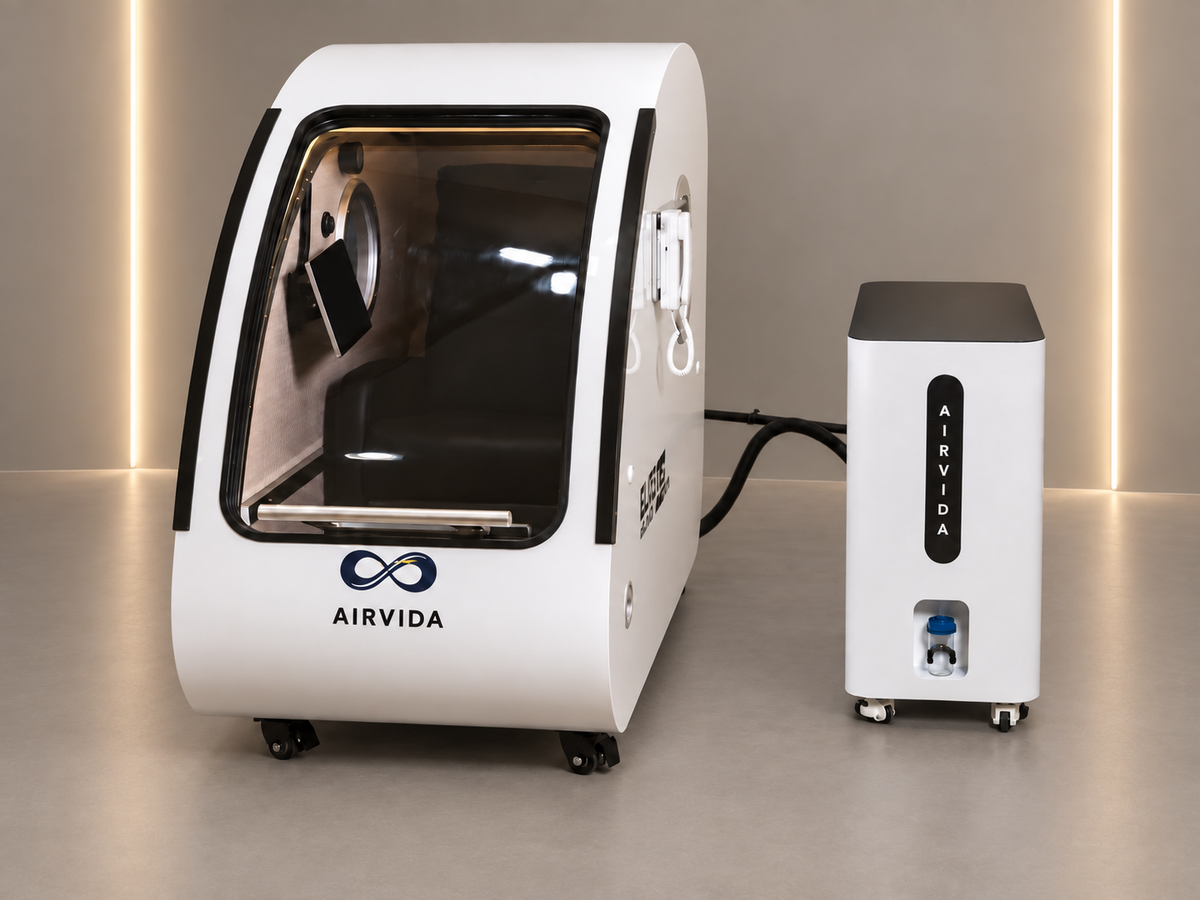 White Airvida hyperbaric chamber with external oxygen system, clear front door, interior seat, control features, and wheels displayed in a modern wellness room.