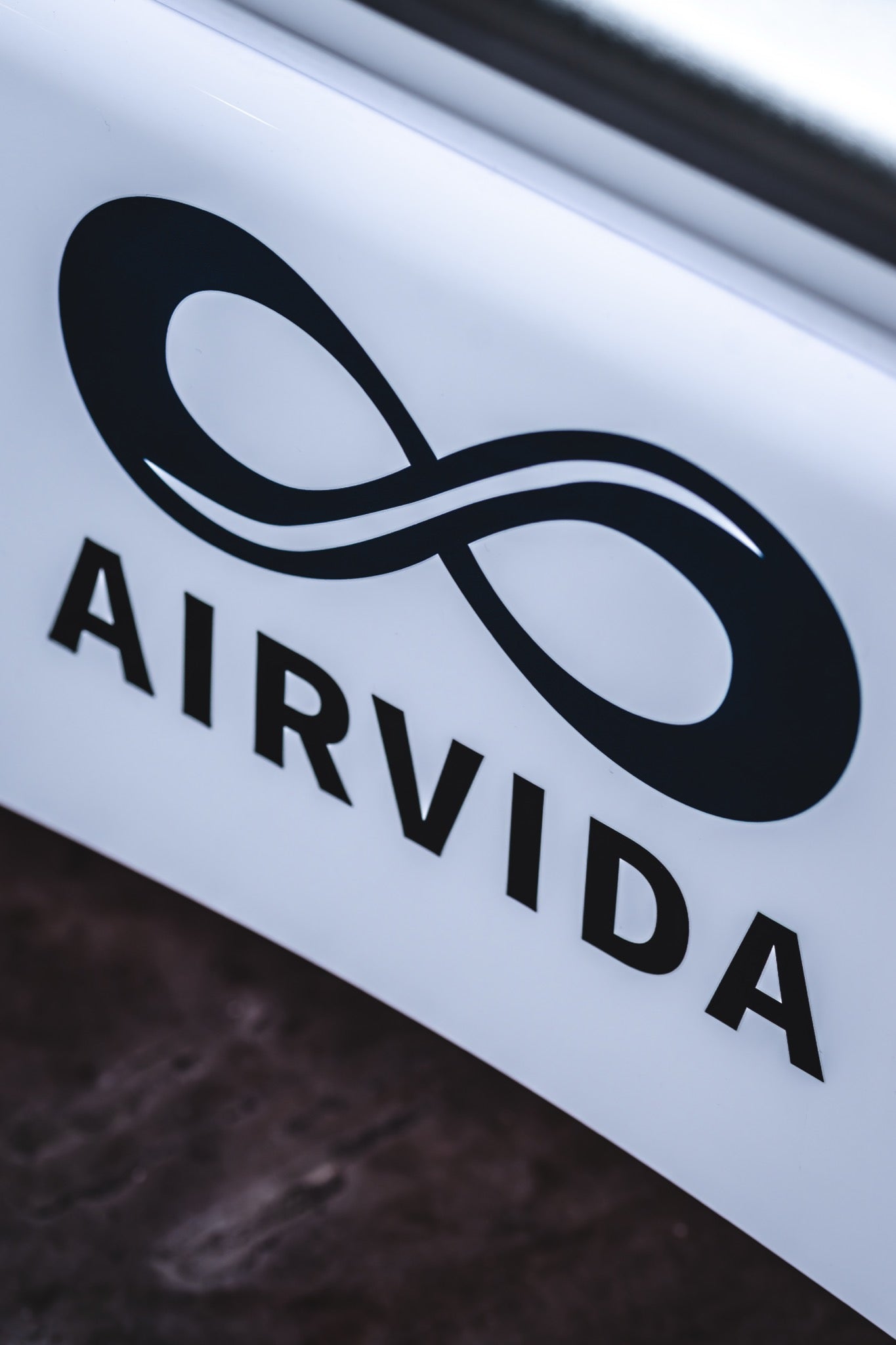 Close-up of the AIRVIDA logo featuring a sleek black infinity symbol and bold lettering on a clean white panel with a dark floor in the foreground.