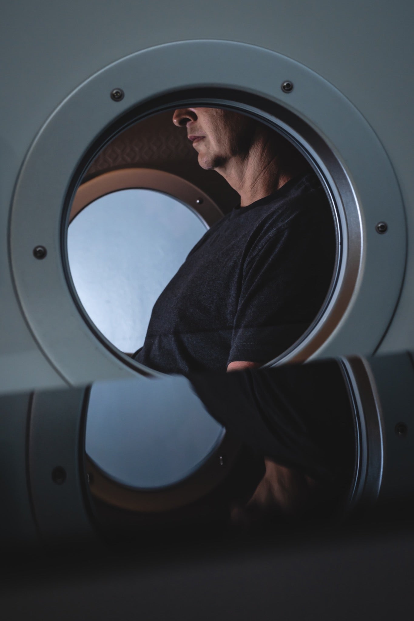 Person seated inside a hard-shell hyperbaric chamber, viewed through its circular side window.