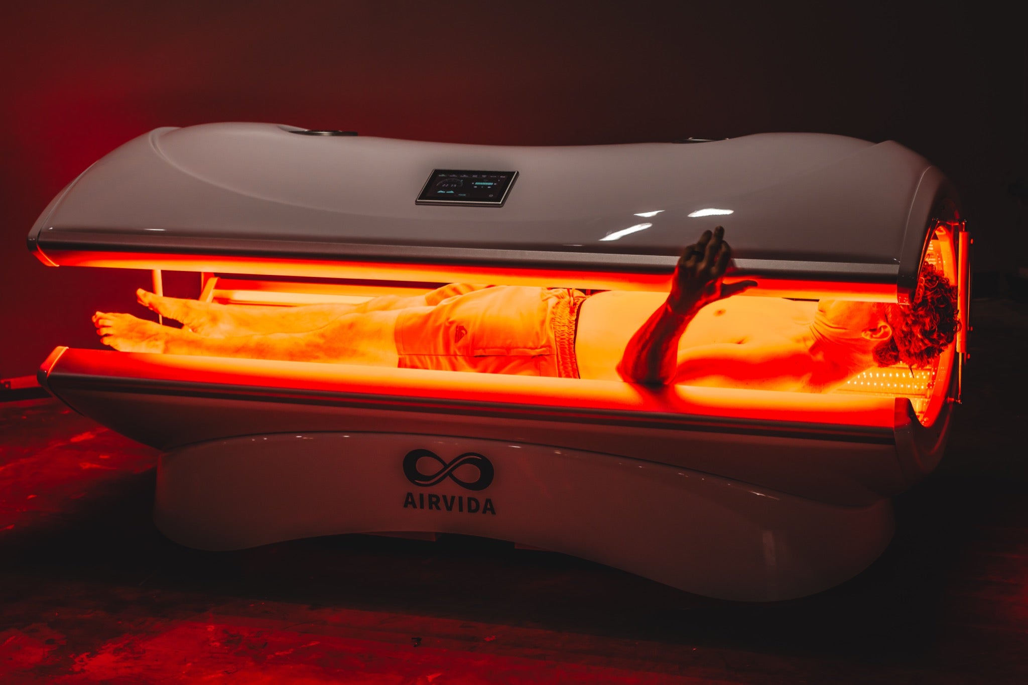 Man relaxing inside an AIRVIDA red light therapy bed, surrounded by bright red LED light for full-body exposure, with a control screen above.