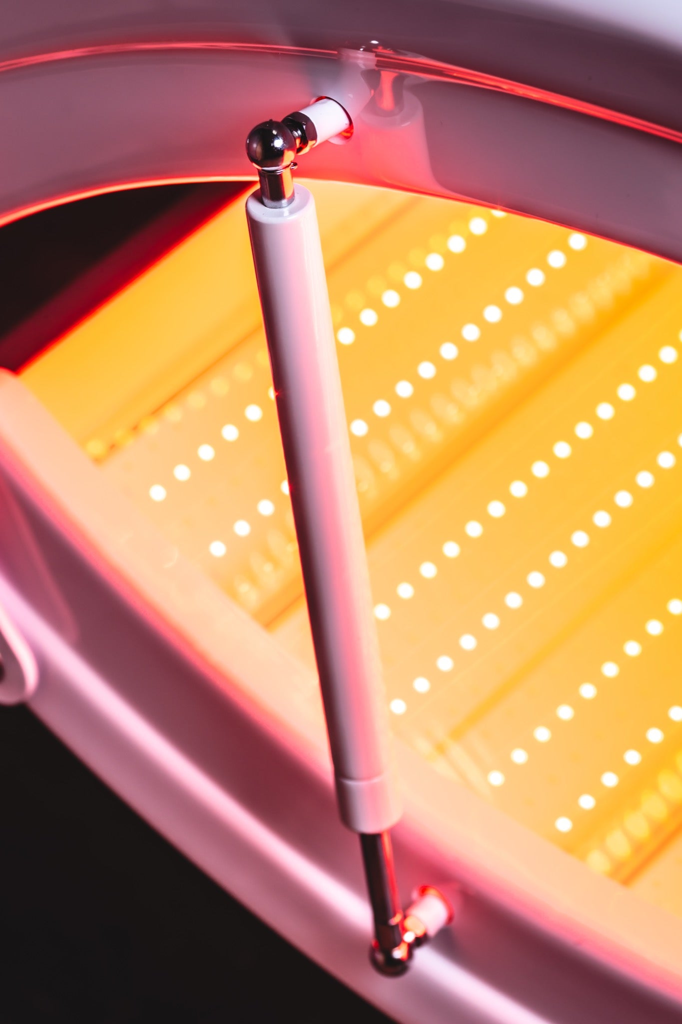 Close-up of a red light therapy bed featuring rows of bright LED lights, a clear curved canopy, and a support arm illuminated by a warm red glow.
