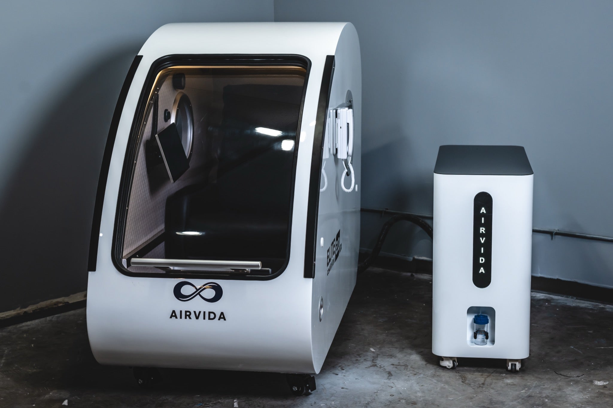 Airvida hard-shell hyperbaric chamber with a matching oxygen system unit, shown together in a modern studio setting.