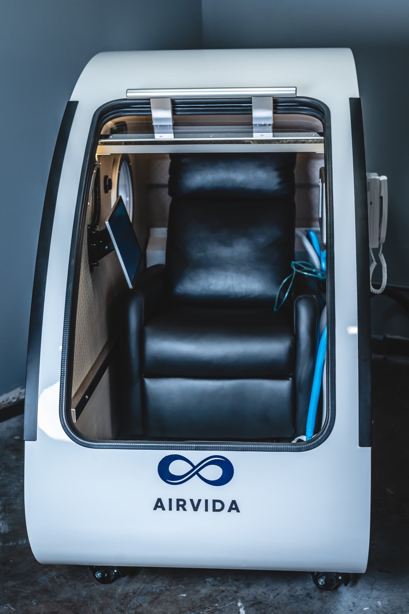 Airvida hard-shell hyperbaric chamber with an open front, showing a reclining seat, display, and oxygen tubing inside.