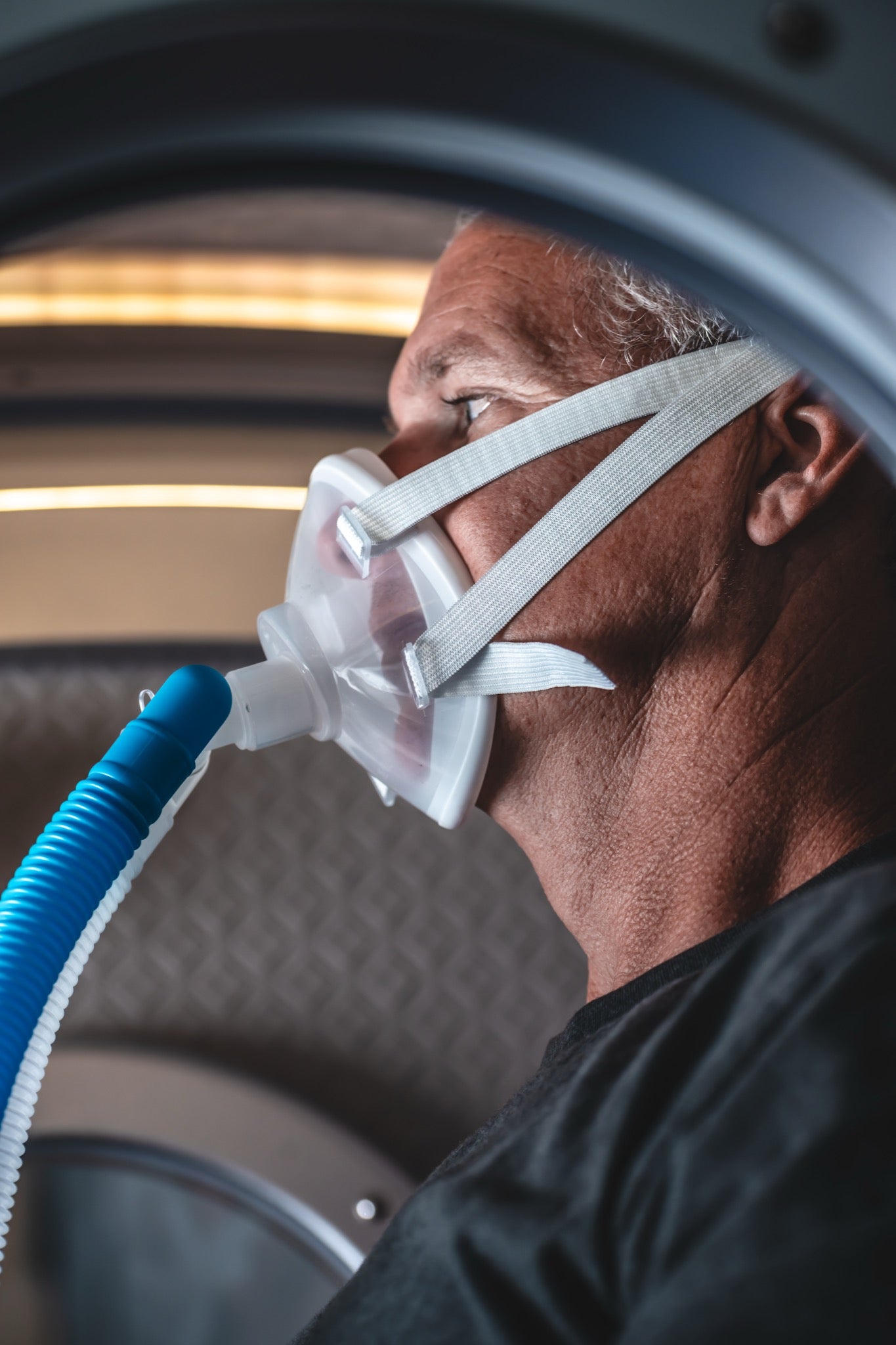 Man wearing an oxygen mask connected to a breathing tube while seated inside a hyperbaric chamber during an HBOT session.