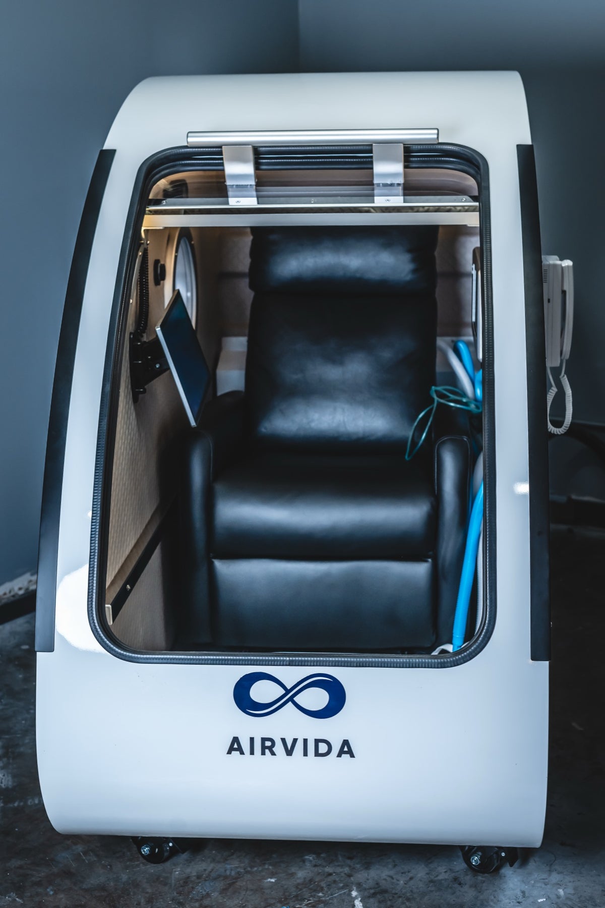 Front view of an Airvida hyperbaric oxygen chamber with an open entry, black reclining seat, interior display, oxygen tubing, and a clear viewing window.