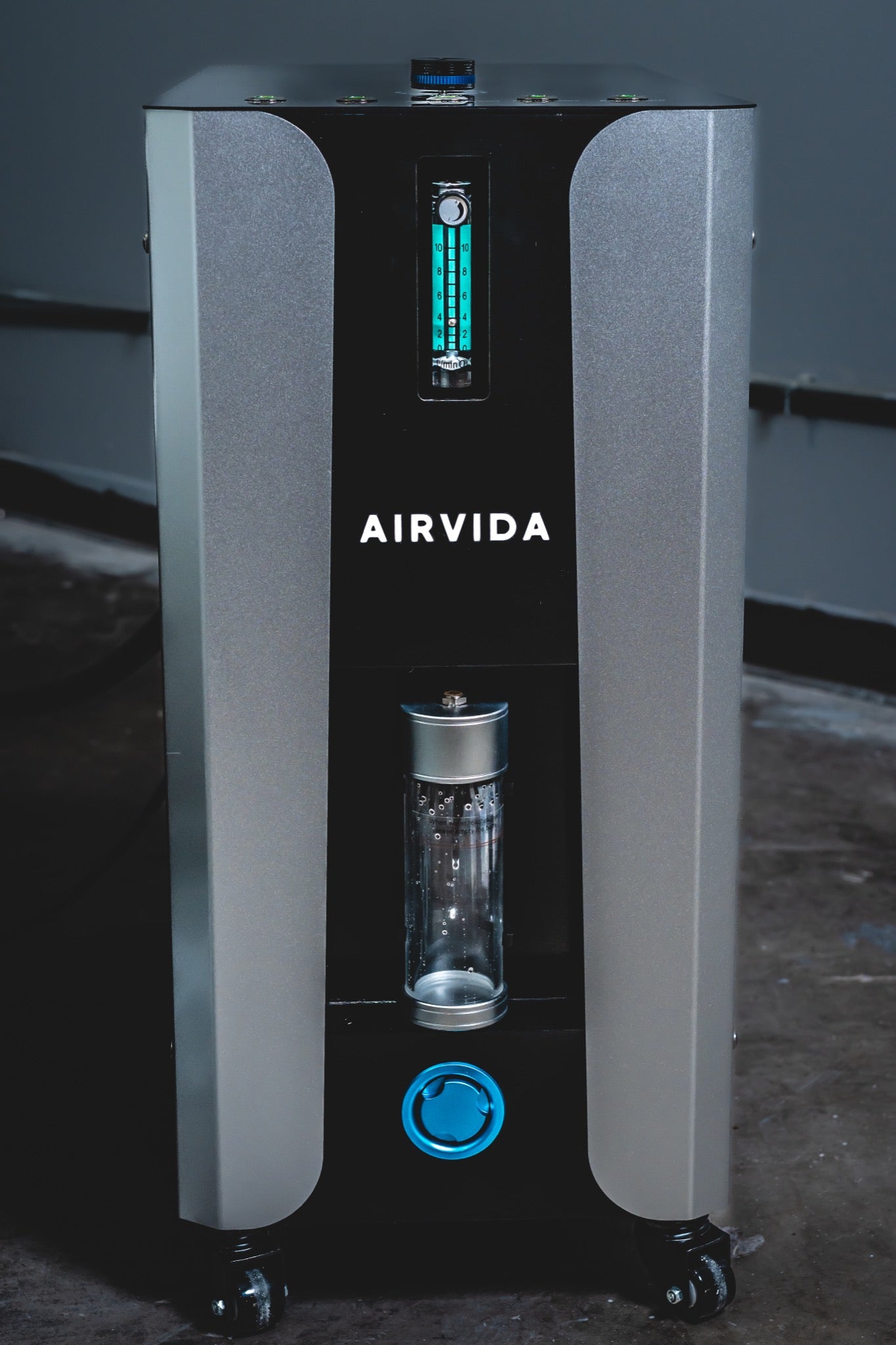 AIRVIDA oxygen concentrator with a sleek black and silver design, featuring a flow meter, humidifier bottle, blue outlet, controls, and wheels.