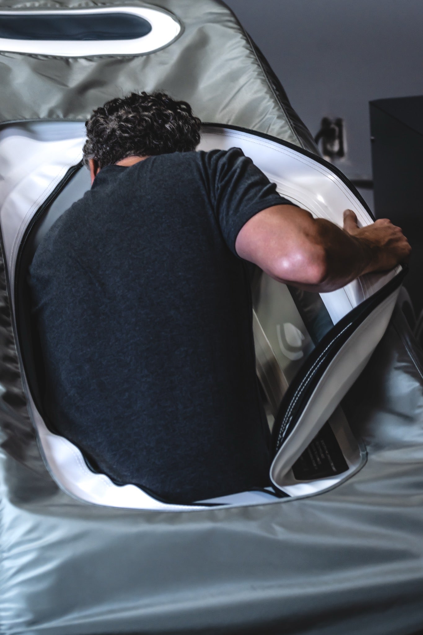 A man in a black t-shirt crawling inside AIRVIDA soft-shell hyperbaric chamber with a clear plastic window.