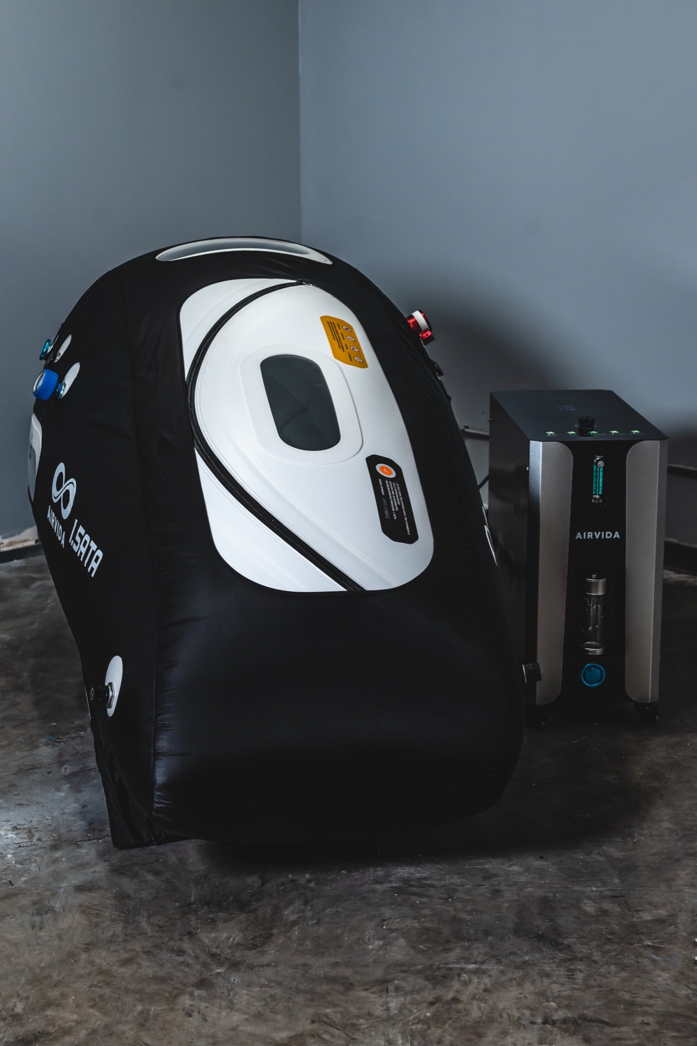 AIRVIDA 1.5 ATA soft-shell hyperbaric chamber with viewing windows and pressure valves, displayed beside its separate oxygen concentrator unit.