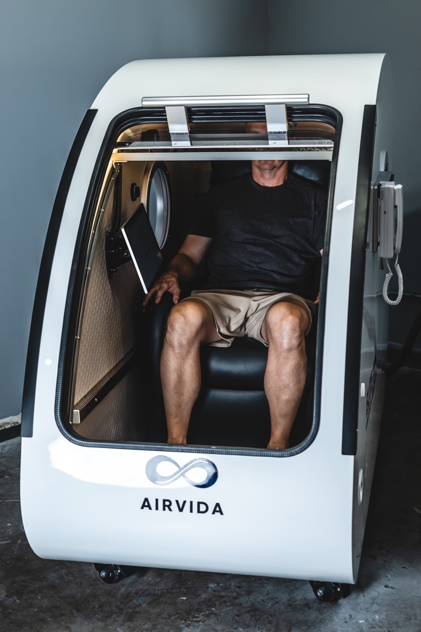 Person seated inside an Airvida hard-shell hyperbaric chamber with the large front door partially open.
