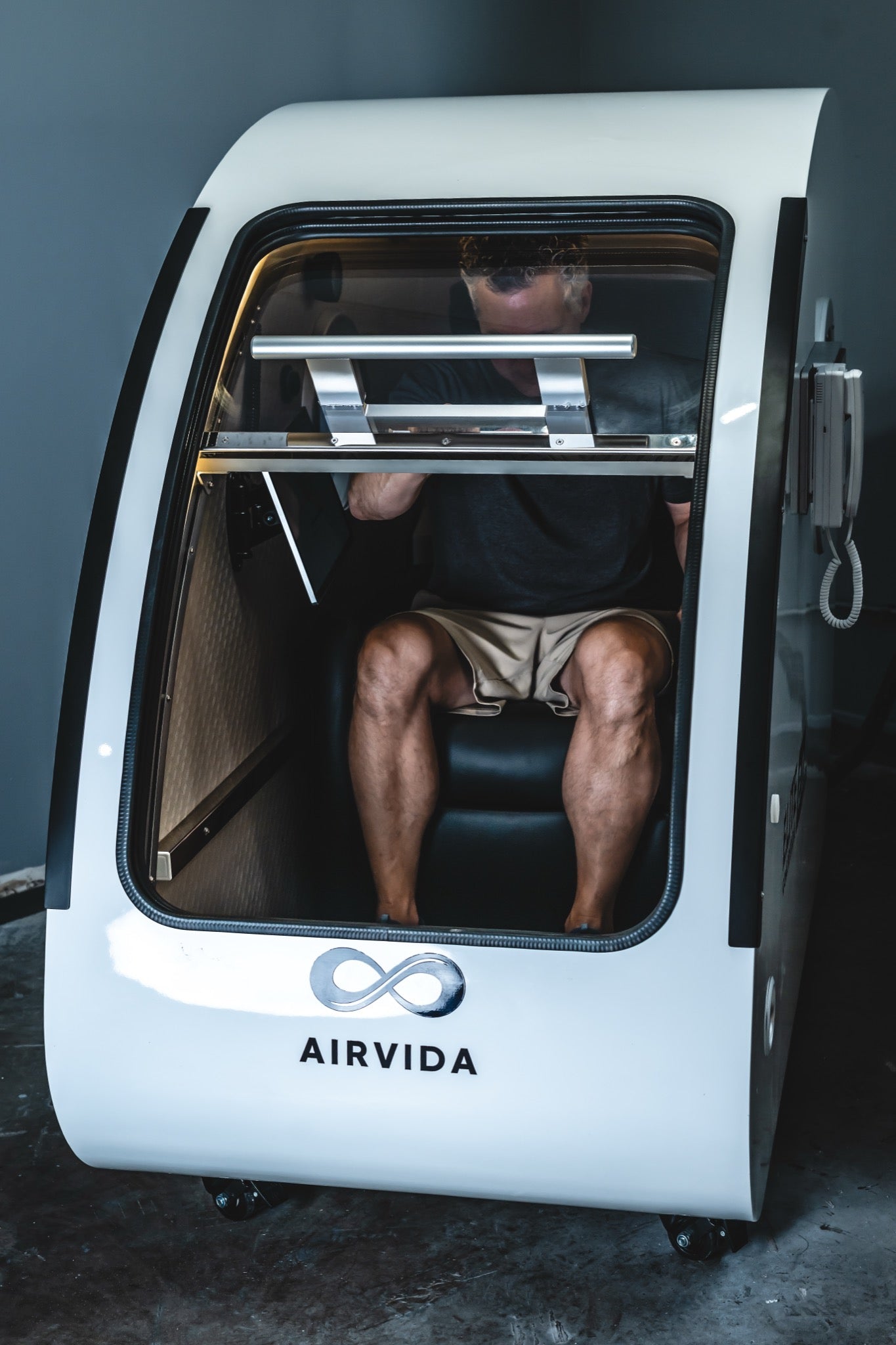 Person seated inside an Airvida hard-shell hyperbaric chamber with a large transparent front door.
