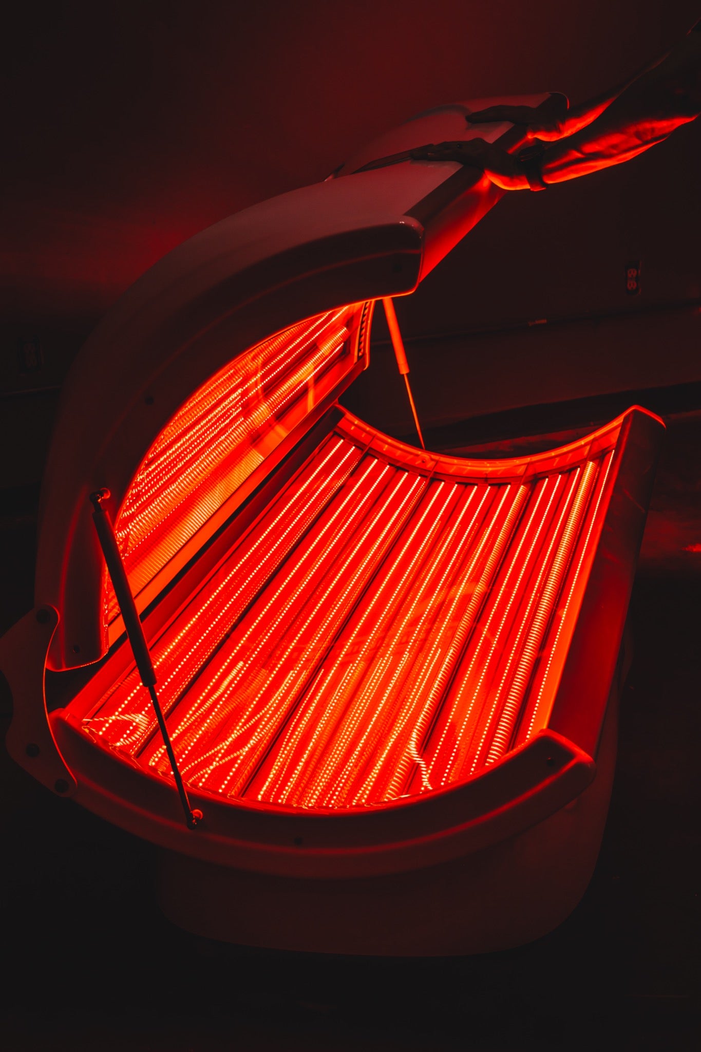 Open AIRVIDA red light therapy bed glowing with rows of bright red LEDs as a person lifts the upper canopy, revealing the full-body treatment area.