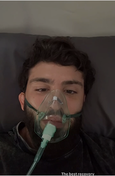 Person lying down wearing a clear oxygen mask with green straps, looking at the camera while receiving respiratory support indoors.