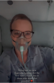 Person wearing glasses and a nasal oxygen mask, reclining on a bed or chair during oxygen or medical therapy.
