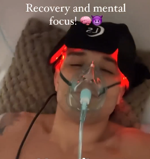 Person lying down wearing an oxygen mask and black cap with red light therapy, eyes closed, resting during a recovery session.