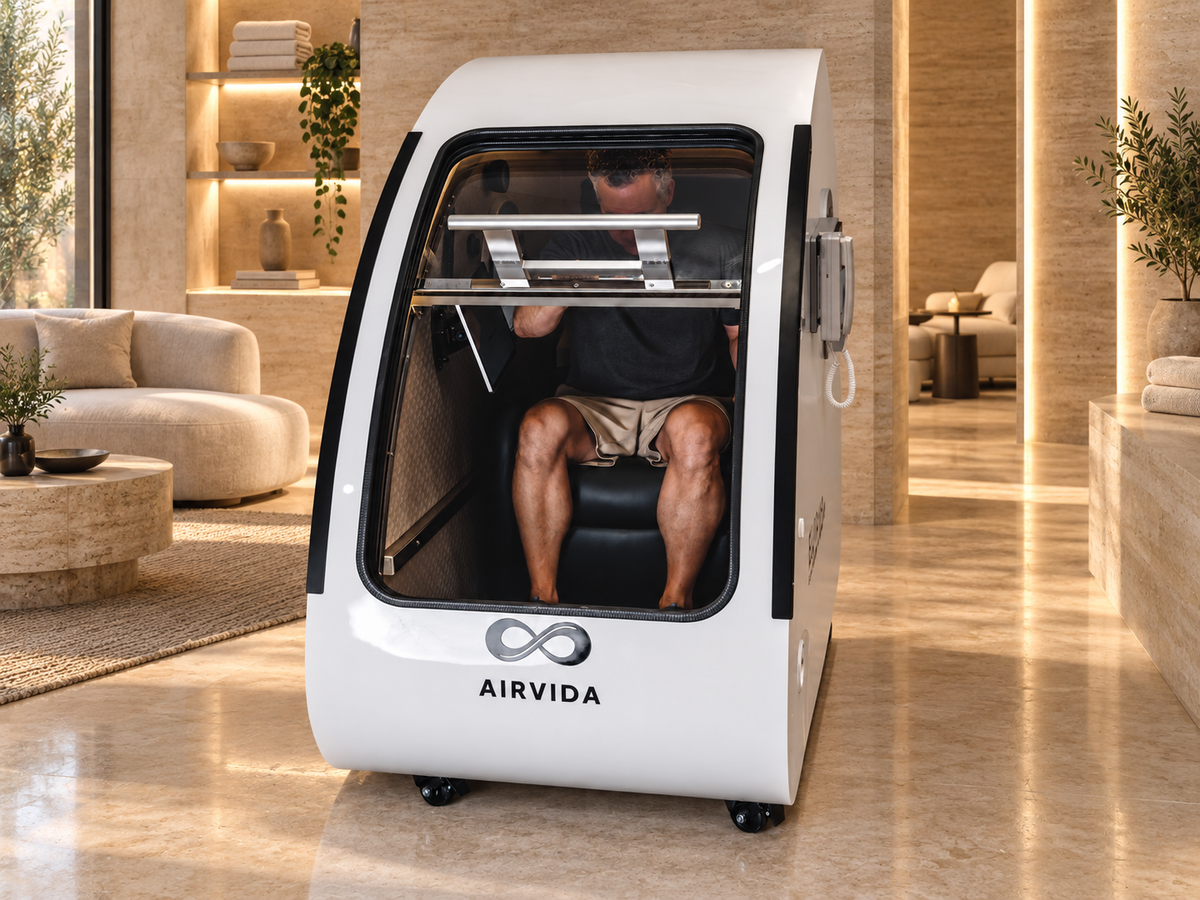 Man seated inside a white Airvida hyperbaric chamber with a clear front door, displayed in a luxurious wellness room with warm lighting, seating, and spa decor.