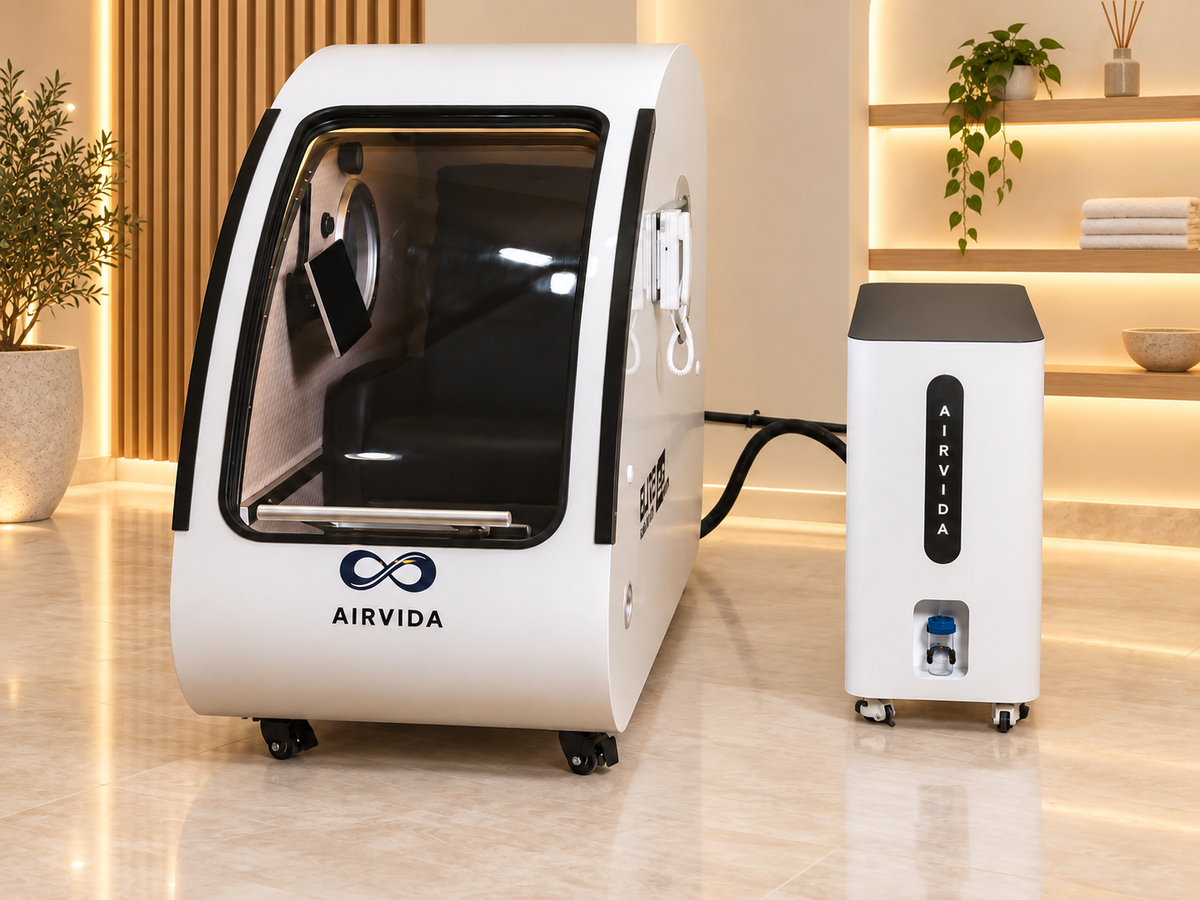 White Airvida hyperbaric chamber with clear entry door and oxygen system displayed in a modern wellness room with warm lighting, wood accents, plants and towels.