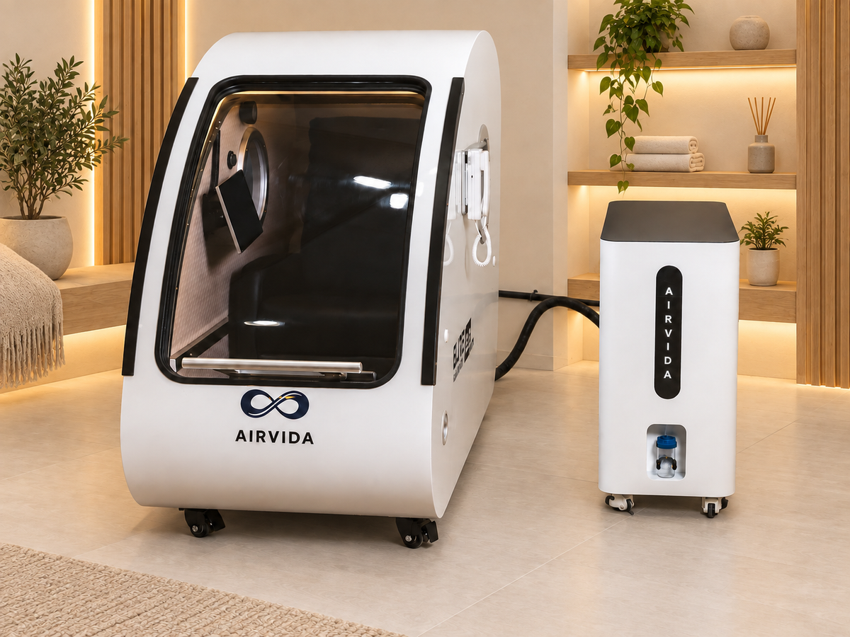 White Airvida hyperbaric chamber with clear entry door and oxygen system displayed in a warm modern wellness room with wood shelves, plants and folded towels.