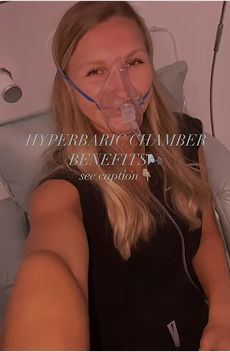 Woman wearing an oxygen mask inside a hyperbaric chamber, taking a selfie while undergoing hyperbaric oxygen therapy for wellness benefits.