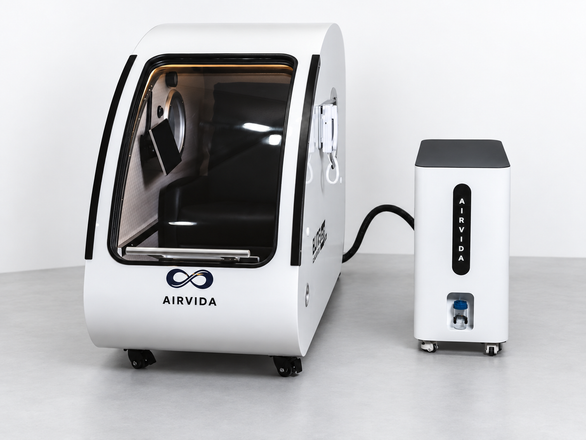 White Airvida hyperbaric chamber with clear curved door and matching oxygen unit connected by a hose, displayed on wheels against a clean white studio backdrop.