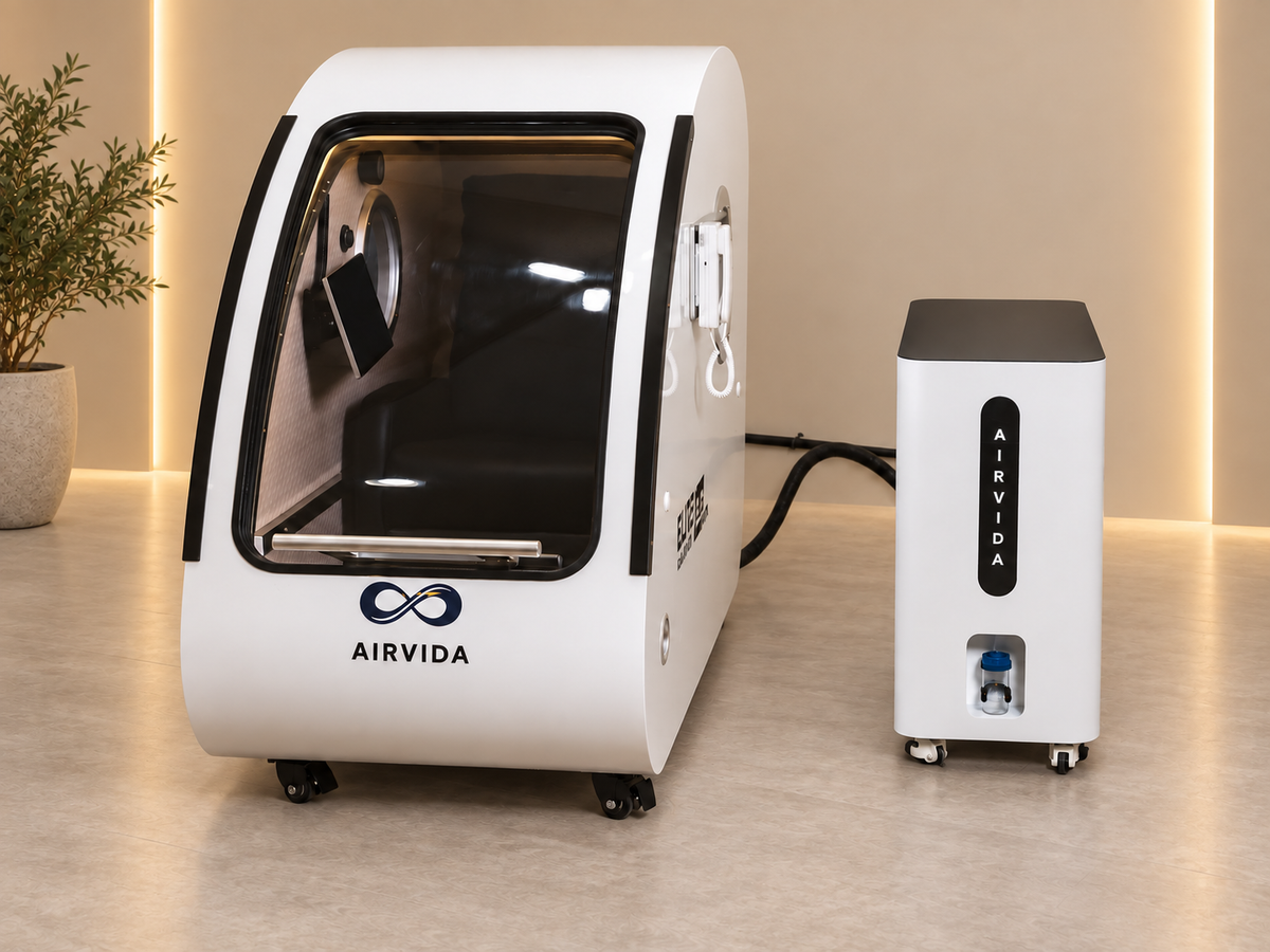 White Airvida hyperbaric chamber with external oxygen system, clear front door, interior seat and wheeled design displayed in a warm modern wellness room.