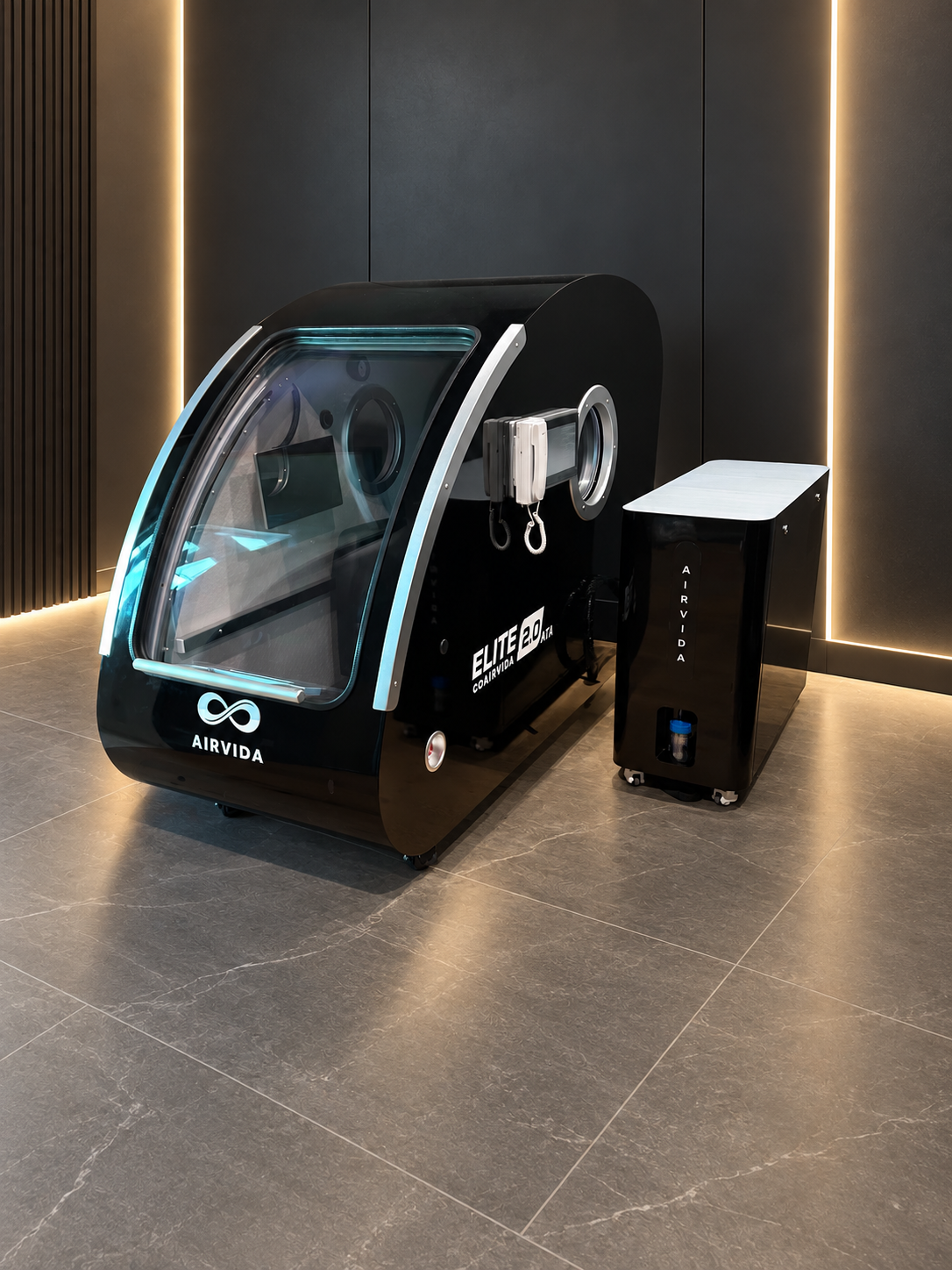Black Airvida Elite 2.0 hyperbaric chamber with external oxygen system in a sleek wellness room featuring dark walls, tiled floors, and warm vertical lighting.