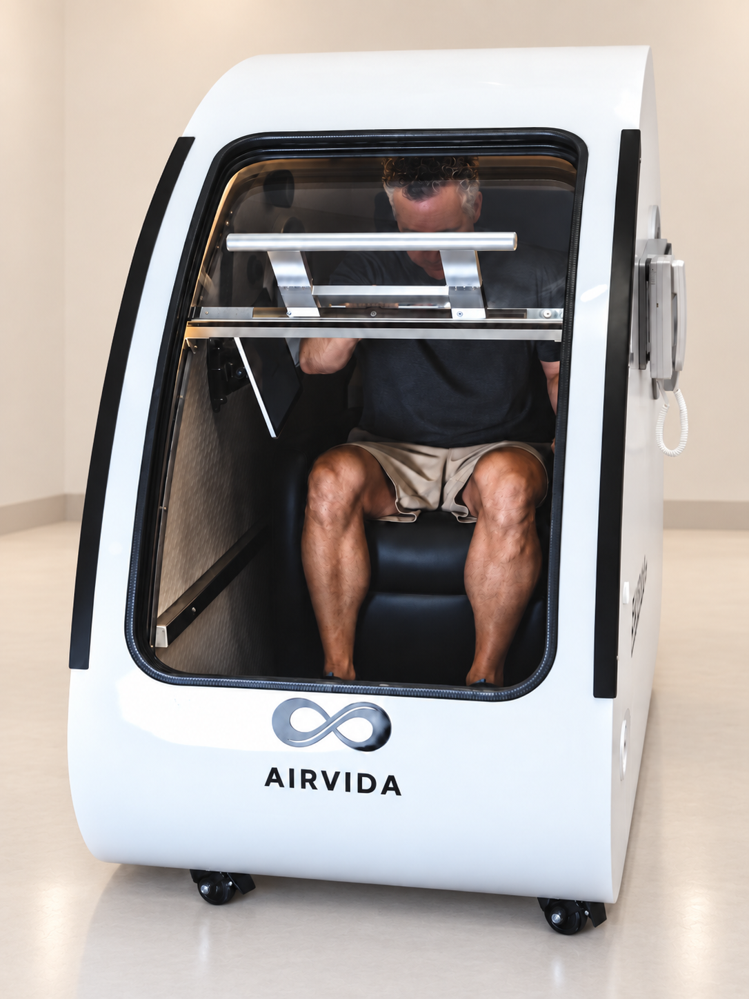 Man seated inside a white Airvida hyperbaric chamber, showing the clear front door, interior seat, safety bars, side controls, and modern chamber design.