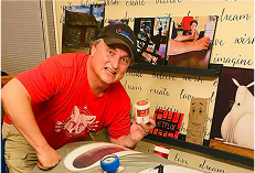 Man in a red shirt and cap posing indoors beside shelves of products, holding a small container and smiling at the camera.