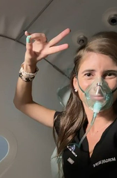 Woman wearing an oxygen mask inside an aircraft cabin, smiling and flashing a peace sign while seated near the window.