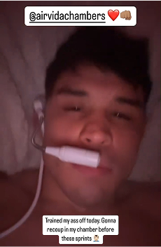 Close-up photo of a young man lying down, wearing earphones, with a white object held between his lips and a relaxed expression.