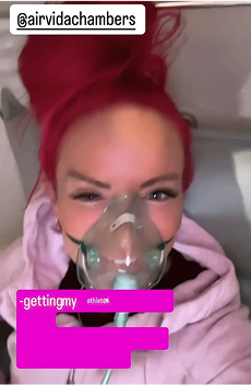 Person wearing a clear oxygen mask and pink hoodie, with red hair tied up, looking at the camera in a clinical or wellness setting.