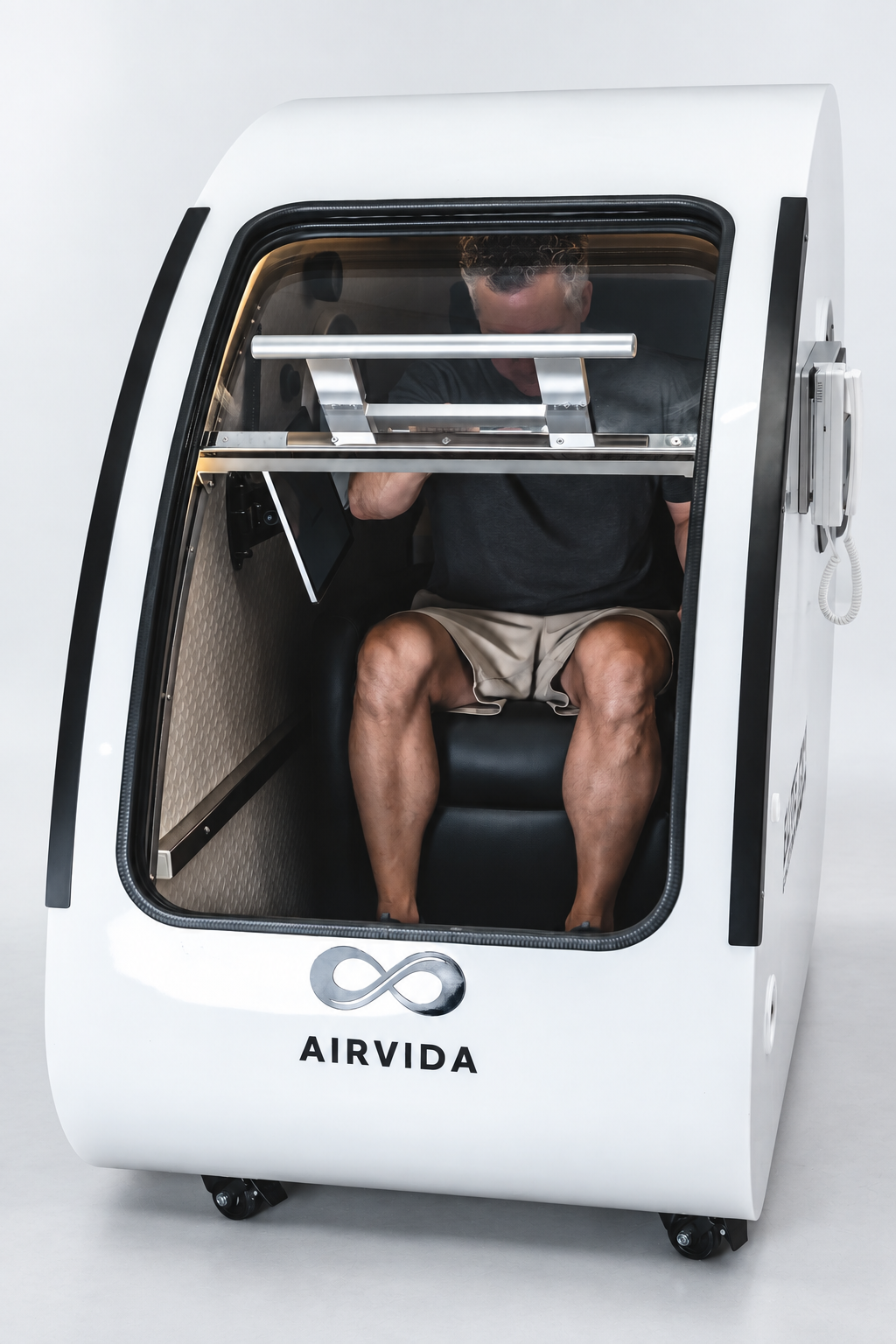 Man seated inside a white Airvida hyperbaric chamber with clear curved door, cushioned seat and metal support bars during a modern wellness therapy session.
